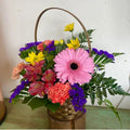 A colorful floral arrangement featuring a variety of flowers such as pink gerberas, yellow daisies, and purple foliage, placed in a woven basket.